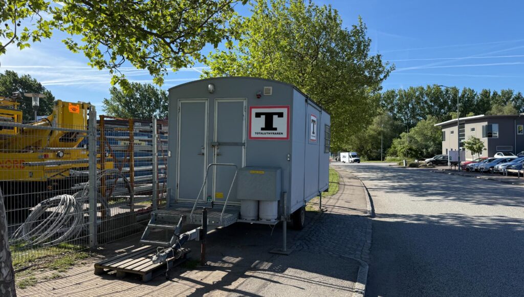 Grå arbetsvagn står vid ett stängsel intill en väg med träd och parkerade bilar i bakgrunden.
