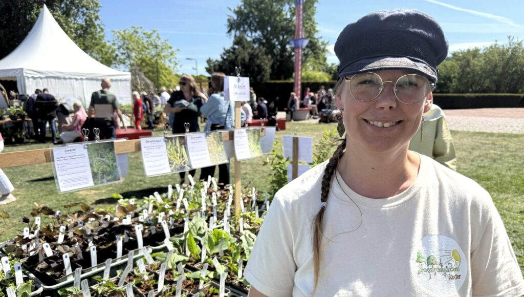 Smidande kvinna i keps och glasögon framför rader av plantor på en utomhusmarknad.