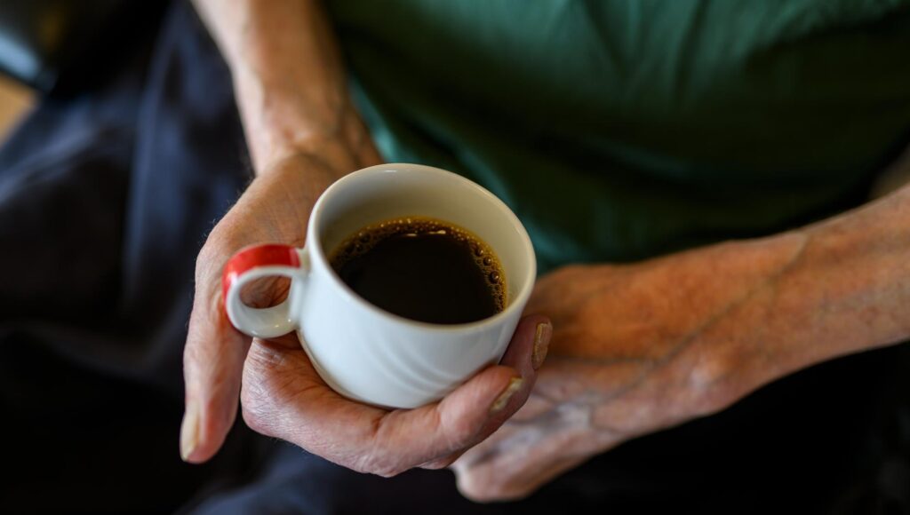 En äldre persons händer håller i en vit kaffekopp med rött öra.