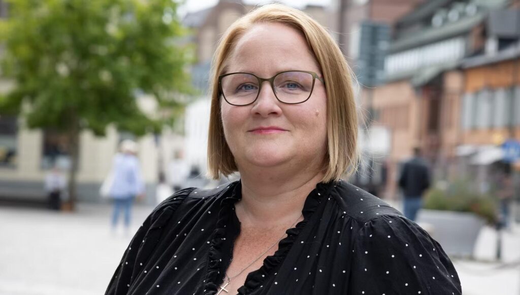 Porträtt av glasögonprydd kvinna i svart prickig blus utomhus i stadsmiljö.