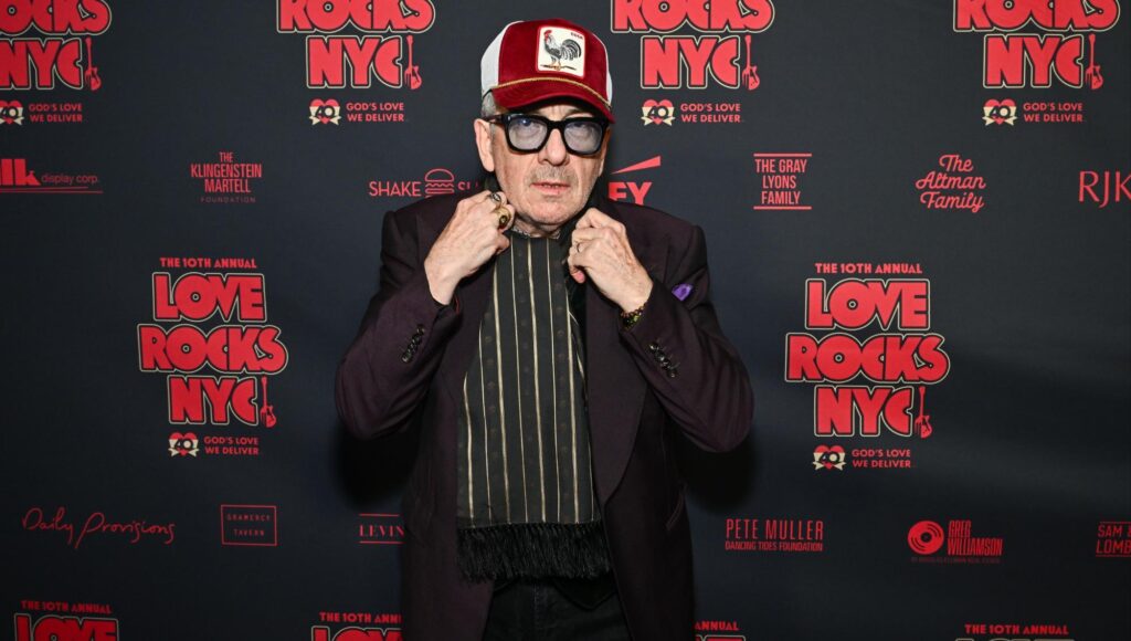 Elvis Costello i lila kavaj, röd keps och mörka glasögon poserar framför en logovägg.