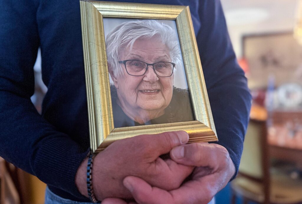 Närbild på en person som håller ett guldinramat porträtt av en leende äldre kvinna med glasögon.
