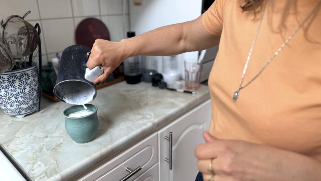 En kvinna häller skummad mjölk från en svart kanna ner i en blågrön keramikmugg i ett kök.