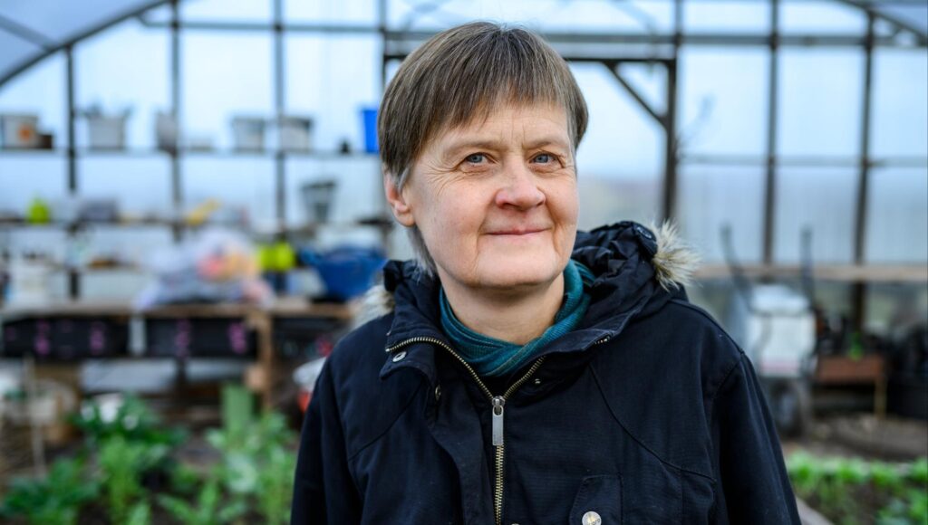 En kvinna i 50-årsåldern i mörk vinterjacka ler mot kameran inuti ett växthus.