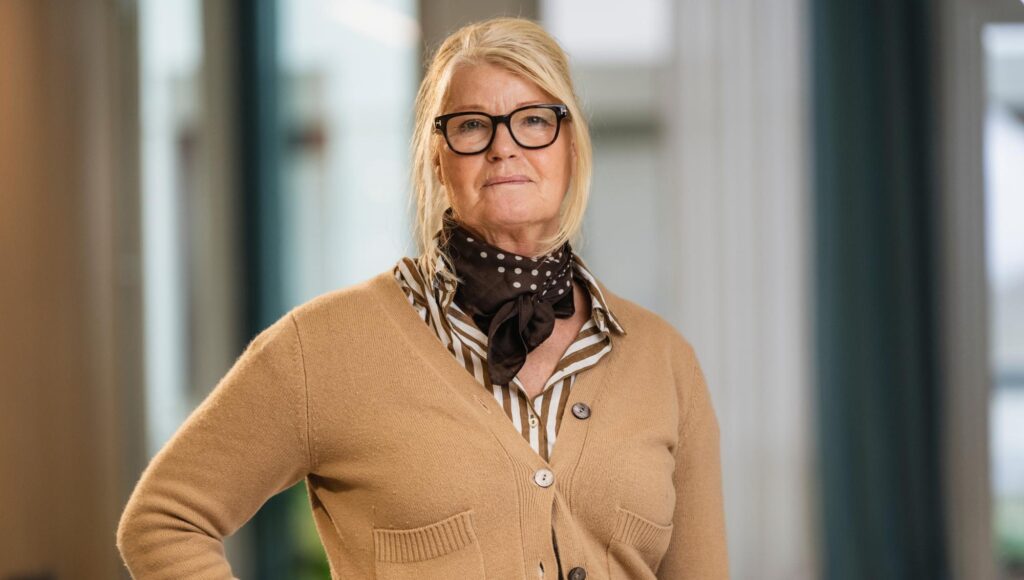 Blond kvinna i 60-årsåldern med glasögon, brun kofta och prickig scarf i ljus inomhusmiljö.