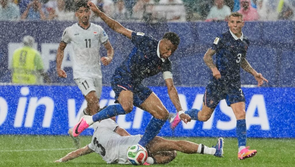 Fotbollsspelare i blå och vit matchdräkt kämpar om bollen i kraftigt regn på en arena.