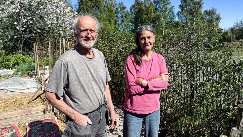 En äldre man och kvinna står i en solig trädgård med odlingslådor och blommande träd i bakgrunden.