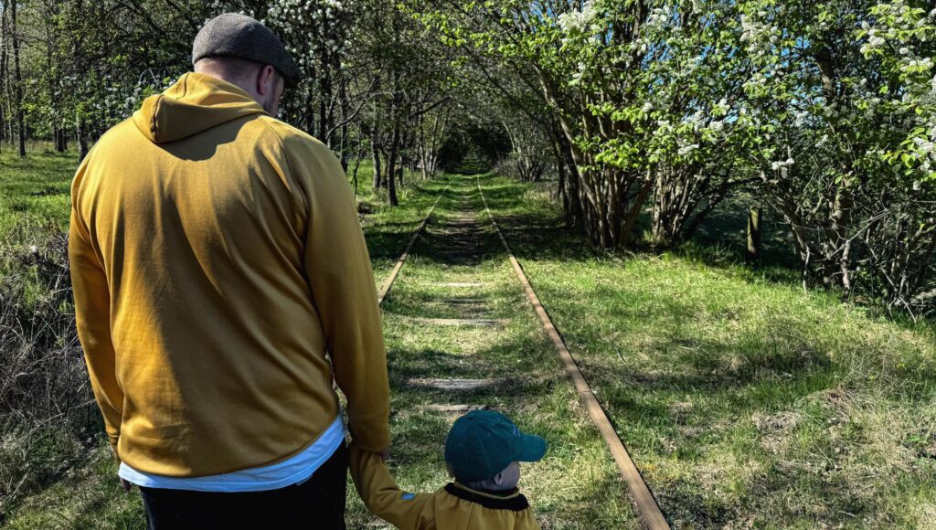 En man och ett barn sedda bakifrån går hand i hand längs ett gammalt tågspår omgivet av blommande träd.