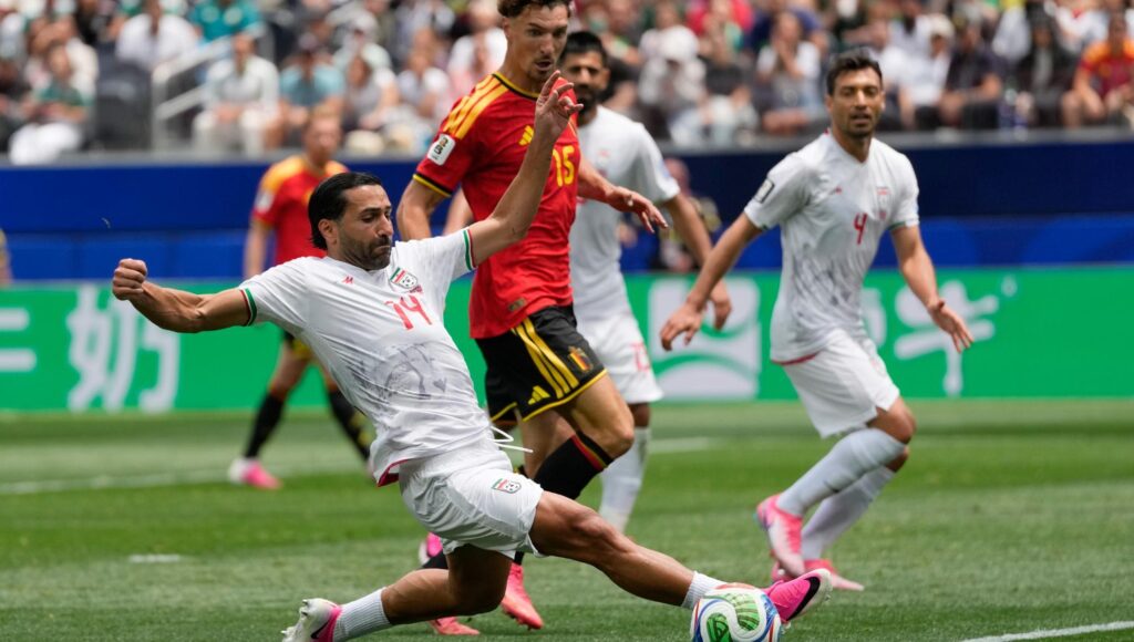 Saman Ghoddos i vita matchkläder glidtacklar bollen framför Thomas Meunier i röd tröja på en gräsplan.