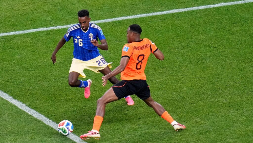Två fotbollsspelare, en i blågul och en i orange matchklädsel, kämpar om bollen på en gräsplan.