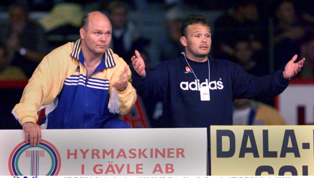 Två män i sportkläder gestikulerar och tittar ut över en plan bakom reklamskyltar.