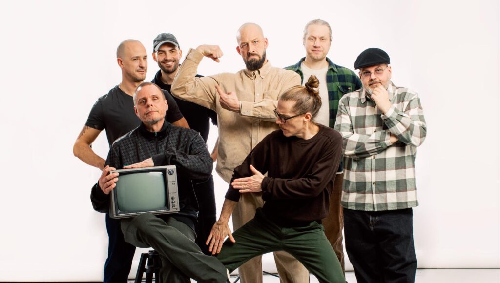 Sju män i en humoristisk gruppostur i en studio, varav en person håller i en gammal tv-apparat.