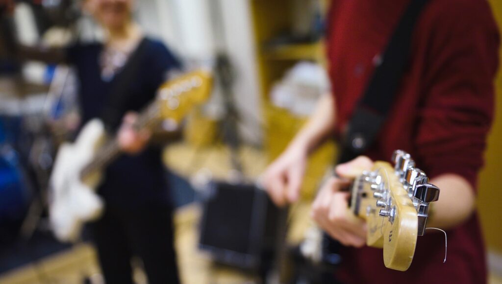 Närbild på gitarrhuvud i fokus medan två personer spelar elgitarr i en replokal.