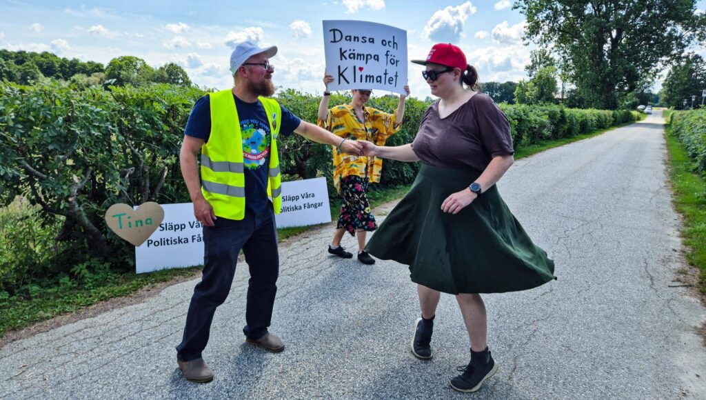 Tre personer dansar och håller upp plakat om klimatet på en asfalterad väg omgiven av grönska.