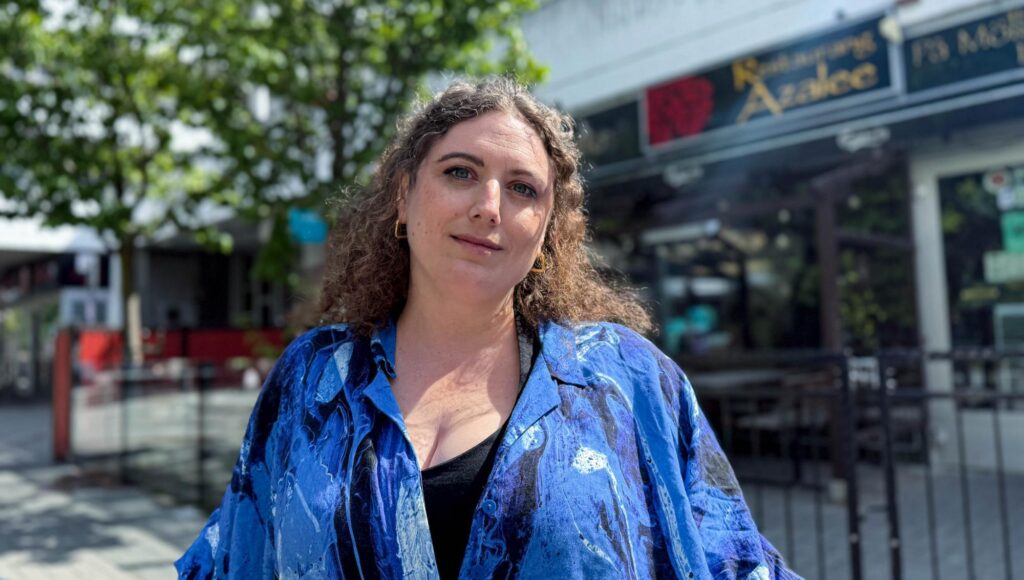 Kvinna med lockigt hår i blåmönstrad skjorta står framför Restaurang Azalee i solljus