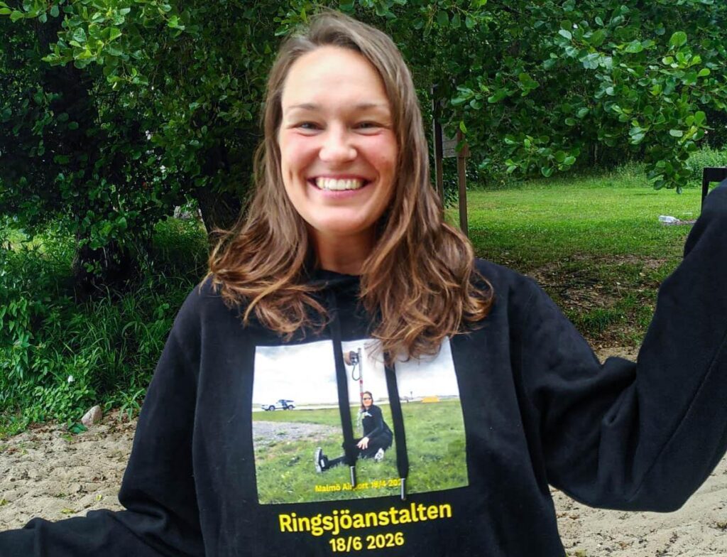 Smidande kvinna i brunett hår bär en svart hoodie med ett tryckt foto och texten Ringsjöanstalten.