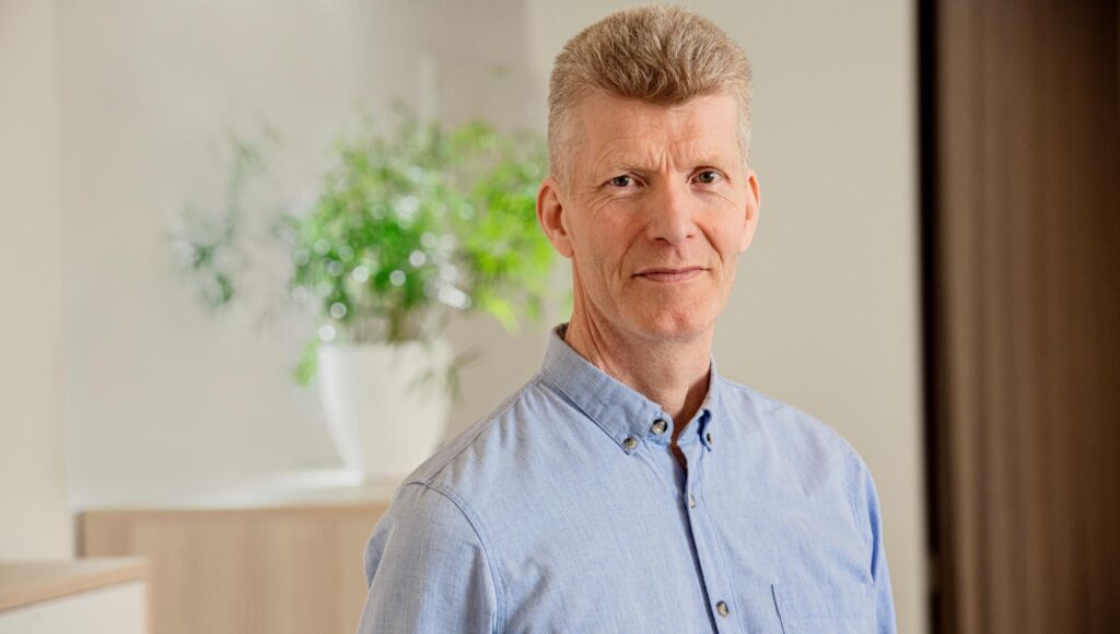 Porträtt på en man med kort ljust hår och blå skjorta framför en grön växt.
