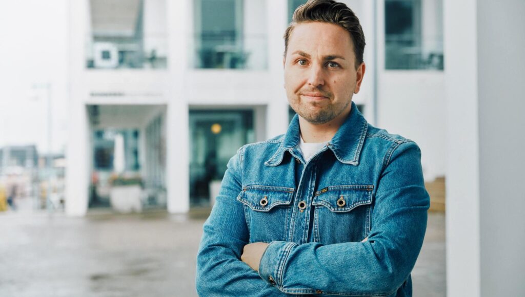 Man i 35-årsåldern med jeansjacka och korslagda armar framför en vit modern byggnad