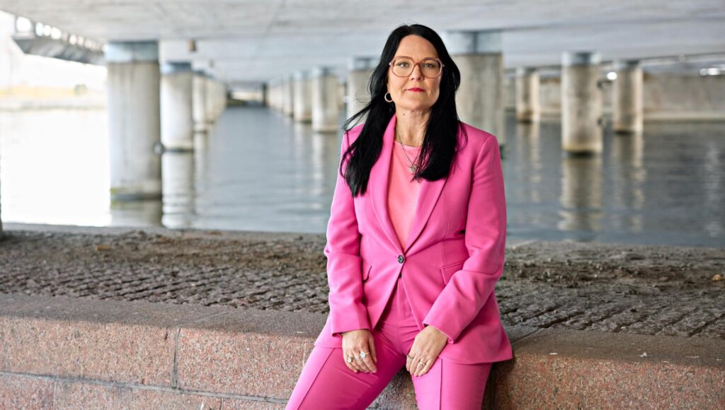 Kvinna med mörkt hår i rosa kostym sitter på en stenkant under en bro vid vatten.