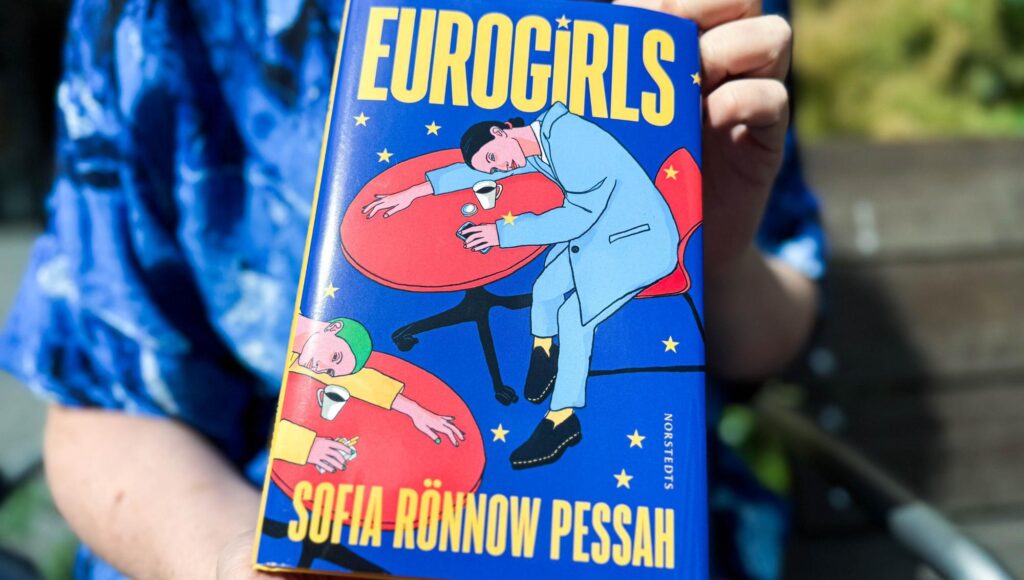 Närbild på boken Eurogirls av Sofia Rönnow Pessah som hålls upp av en person i blå mönstrad tröja.