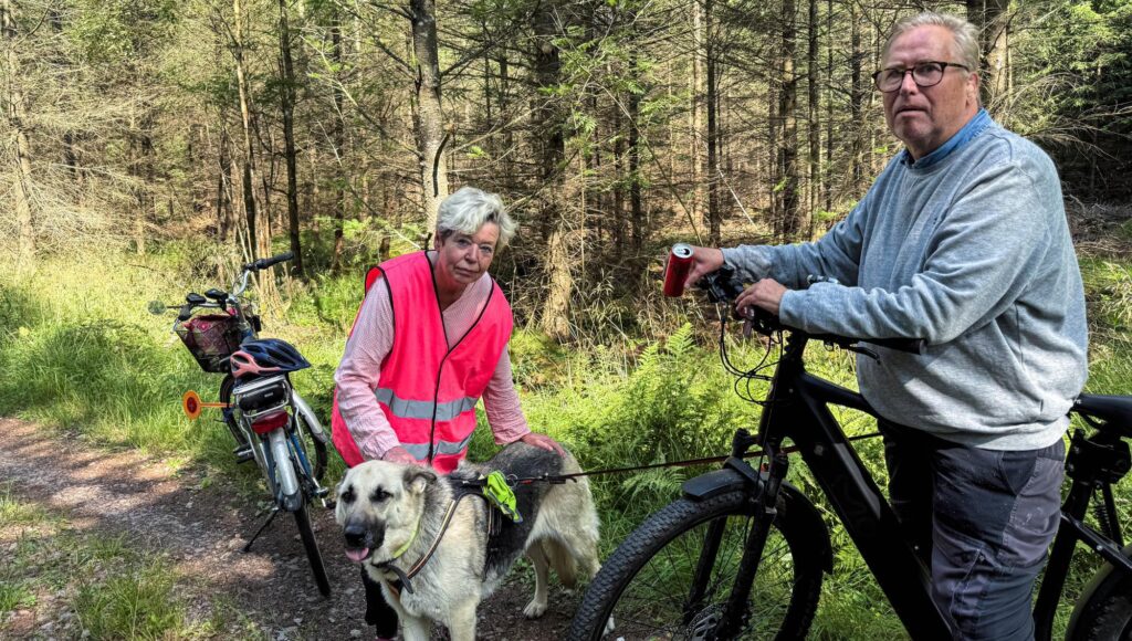 En kvinna i rosa reflexväst och en man med glasögon står vid sina cyklar och en stor hund på en skogsstig.
