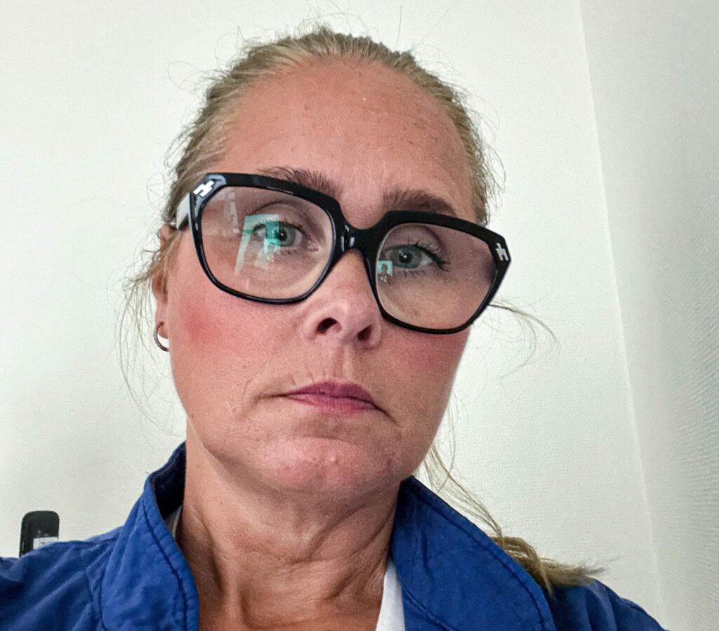 Närbild på kvinna med kraftiga svarta glasögon och blå jacka mot en vit vägg