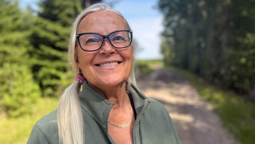 Leende äldre kvinna med glasögon och blont hår uppsatt i hästsvans utomhus på en skogsväg.