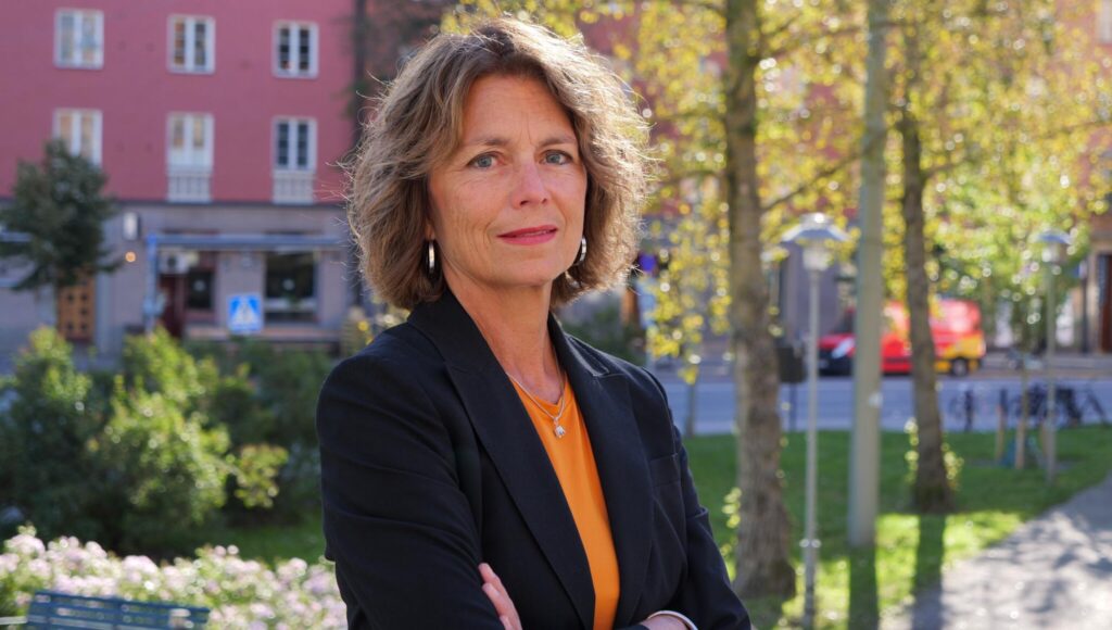 Porträtt av kvinna i mörk kavaj och orange tröja utomhus med grönskande träd och hus i bakgrunden