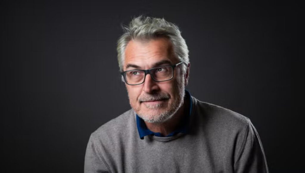 Gråhårig man i glasögon och tröja tittar åt sidan mot en mörk bakgrund.