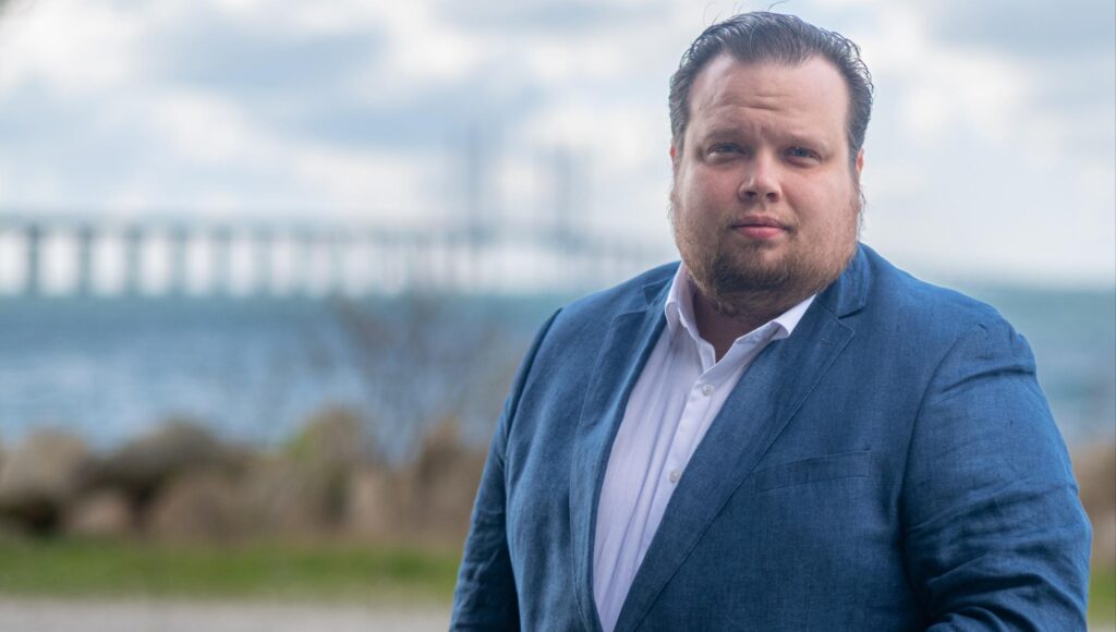 Porträtt av skäggig man i blå kavaj och vit skjorta utomhus med en bro i bakgrunden.