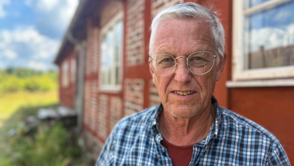 Närbild på en äldre man med glasögon och rutig skjorta framför ett rött korsvirkeshus.