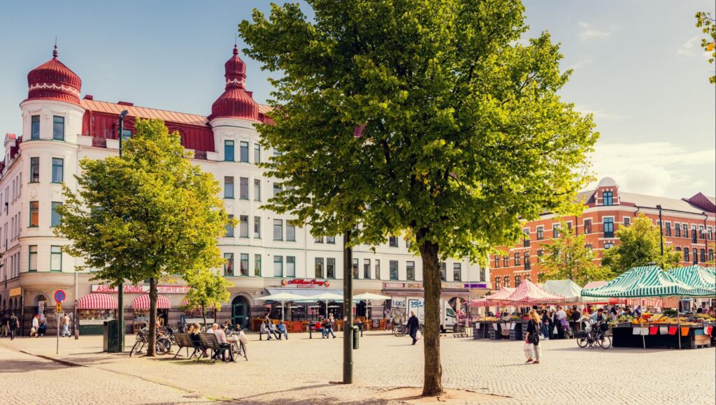 Soligt torg med marknadsstånd, träd och äldre stenhus med röda takkupoler i bakgrunden.