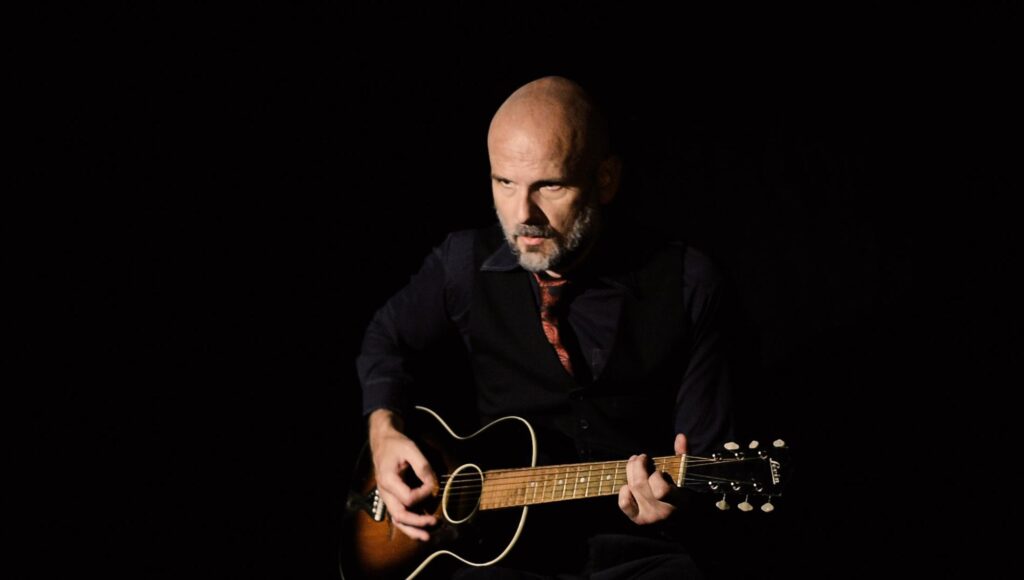 Skallig man i 40-årsåldern med skägg spelar gitarr mot svart bakgrund.