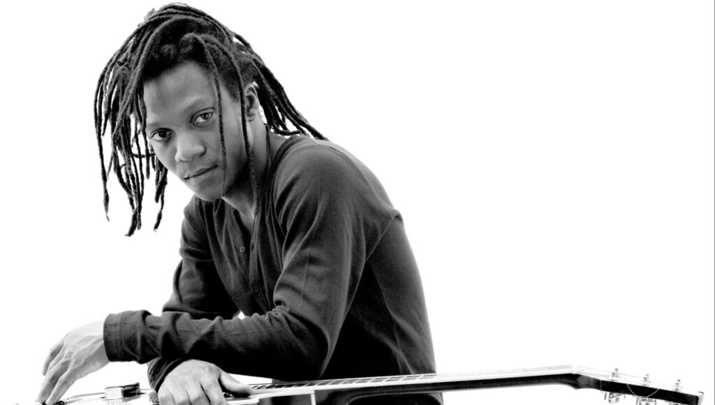 Svartvit bild på man med dreadlocks som lutar sig över en gitarr mot vit bakgrund