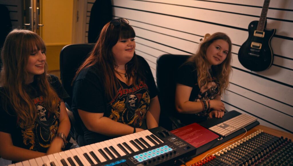 Tre unga kvinnor vid ett mixerbord och klaviatur i en musikstudio med en elgitarr på väggen.