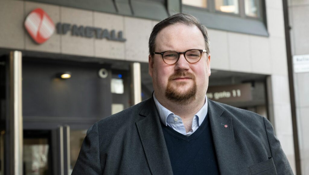 Porträtt av Simon Petersson i glasögon och kavaj framför IF Metalls logotyp och entré.