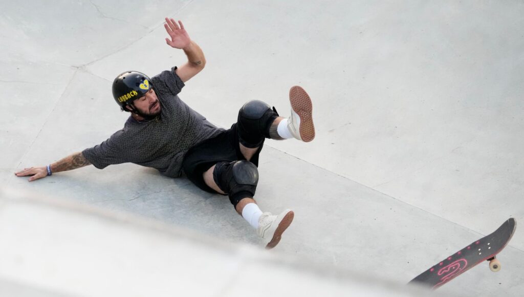 Skateboardåkare i hjälm och knäskydd faller på betongen i en ramp medan brädan rullar iväg.