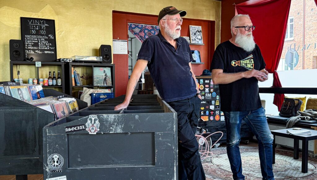 Två äldre män med skägg står i en skivbutik med vinylbackar, ölflaskor och en griffeltavla i bakgrunden.