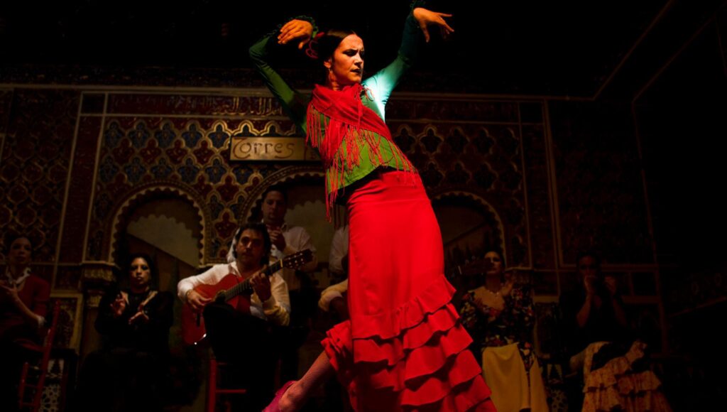 Kvinna i röd flamencoklänning dansar på en scen framför musiker i en dekorativ lokal.
