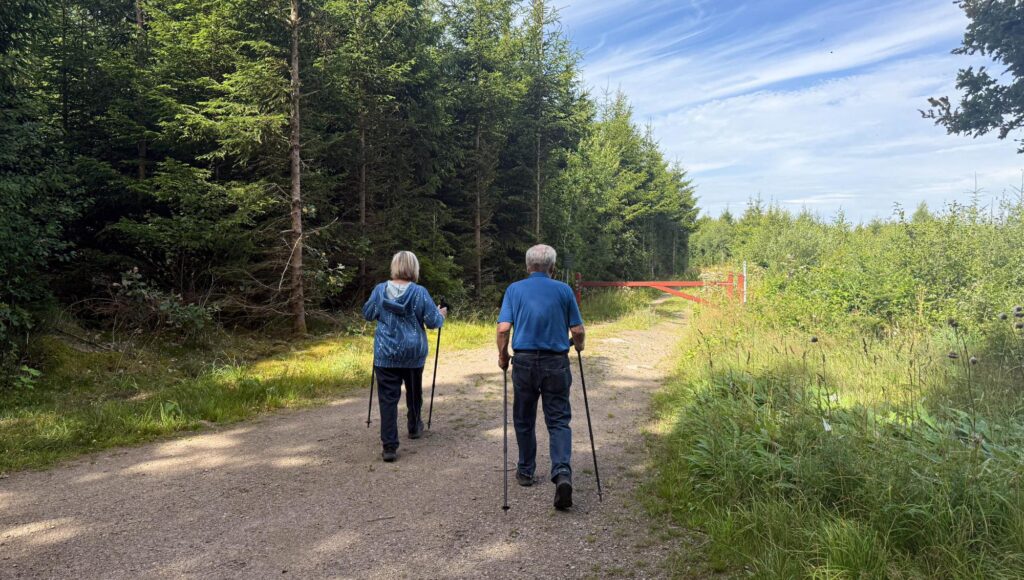 Ett äldre par går med gångstavar bortåt på en grusväg kantad av tät granskog.
