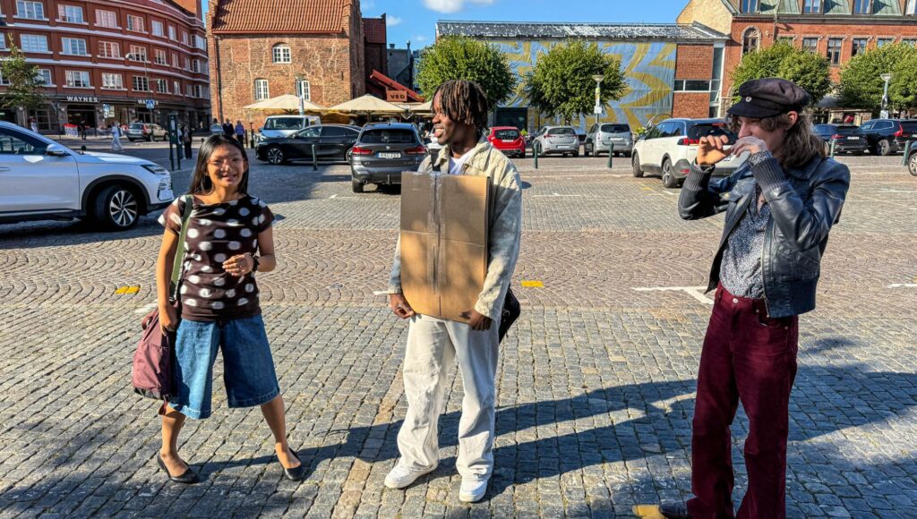 Tre ungdomar står på ett stenlagt torg i solljus, en av dem bär på en stor kartongskiva.