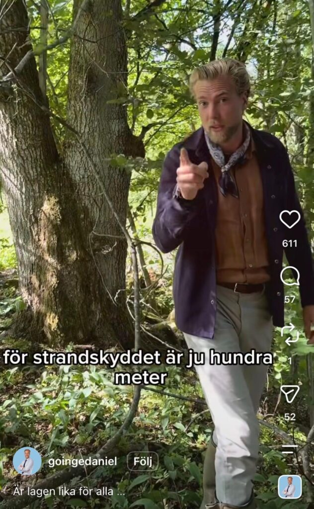 Blond man i skogsmiljö pekar mot kameran och bär en mörk jacka med scarf.