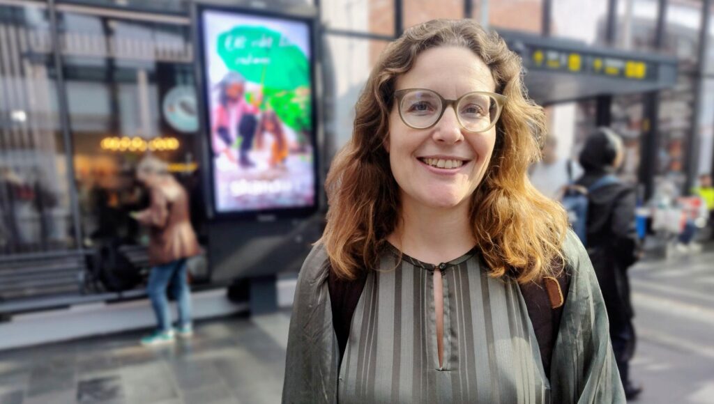 Leende kvinna med glasögon och lockigt hår står framför en digital reklamskärm i en stationsmiljö.