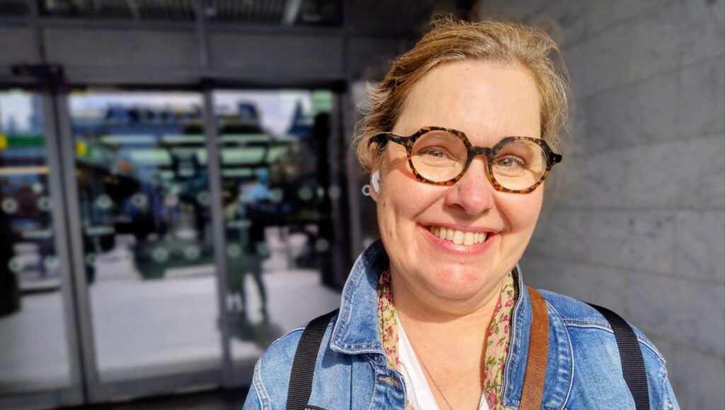 Leende kvinna i glasögon och jeansjacka utomhus i solljus.