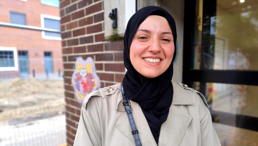 Leende kvinna i svart hijab och beige trenchcoat står framför en tegelvägg utomhus.