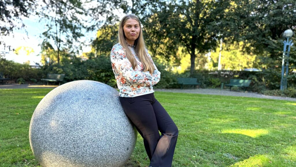 Långhårig kvinna i blommig blus lutar sig mot en stor stenboll i en solig parkmiljö.