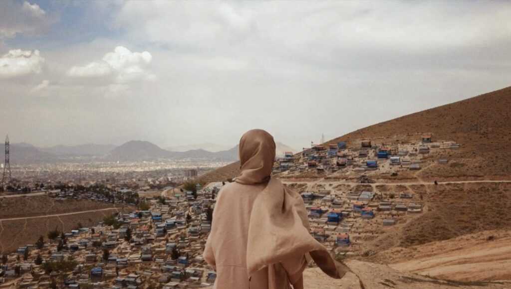 Kvinna i hijab sedd bakifrån blickar ut över en stad av små enkla hus i en bergssluttning under en molnig himmel.