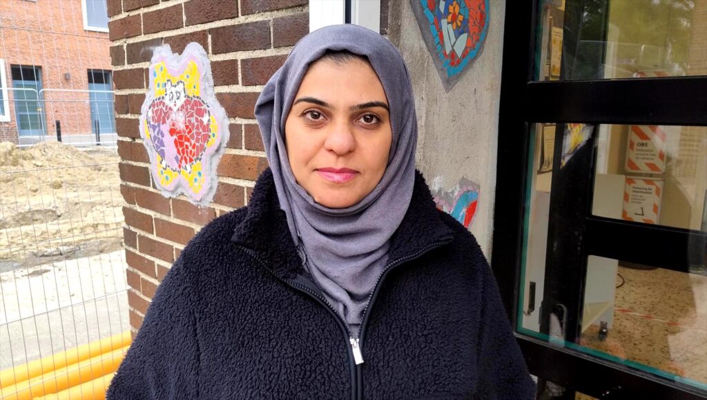 Kvinna i lila hijab och svart fleecejacka står utomhus framför en tegelvägg med mosaikkonst.