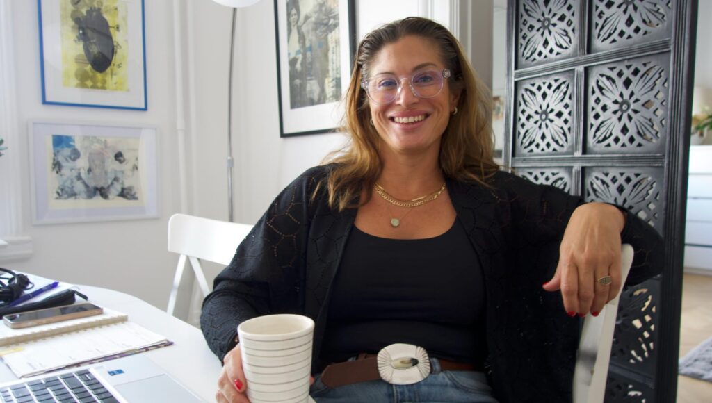 Leende kvinna i 40-årsåldern med glasögon sitter vid ett vitt bord med en kaffekopp framför en laptop.