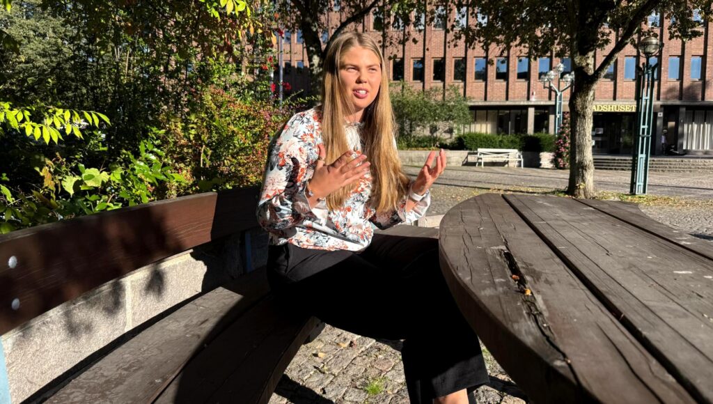 Långhårig kvinna i blommig blus sitter vid ett träbord utomhus och gestikulerar med händerna.